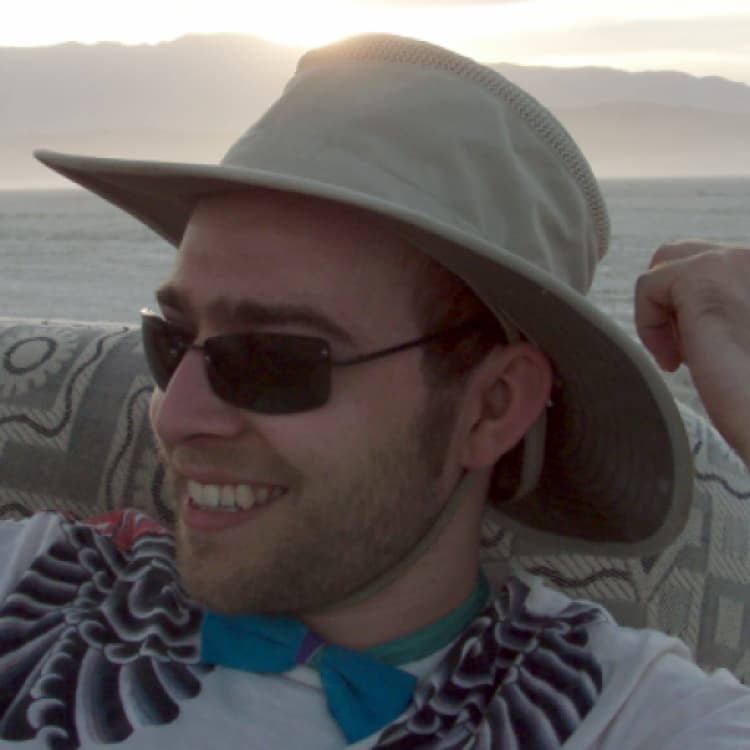 A picture of an unshaved man in a hat wearing a blue bowtie with the sun behind him